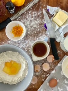 Easter bread ingredients - flour in a large bowl with 2 broken eggs in the centre, maple syrup in a measuring cup, grated zest of an orange in a bowl, block of butter on foil wrapping with a knife at the side of it, milk in a jug and a bowl of butter just partly cut out of the shot and the surface covered in flour with egg shells to the side of the bowl - Pure Maple