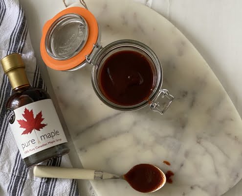 pot of prepared Maple Barbecue Sauce next to bottle of maple syrup, spoon on table covered in sauce