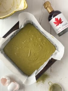 Matcha brownie mix in a lined tin with mixing bowl and maple syrup bottle at the top of shot and egg shells and sieve with matcha powder at the bottom -Pure Maple