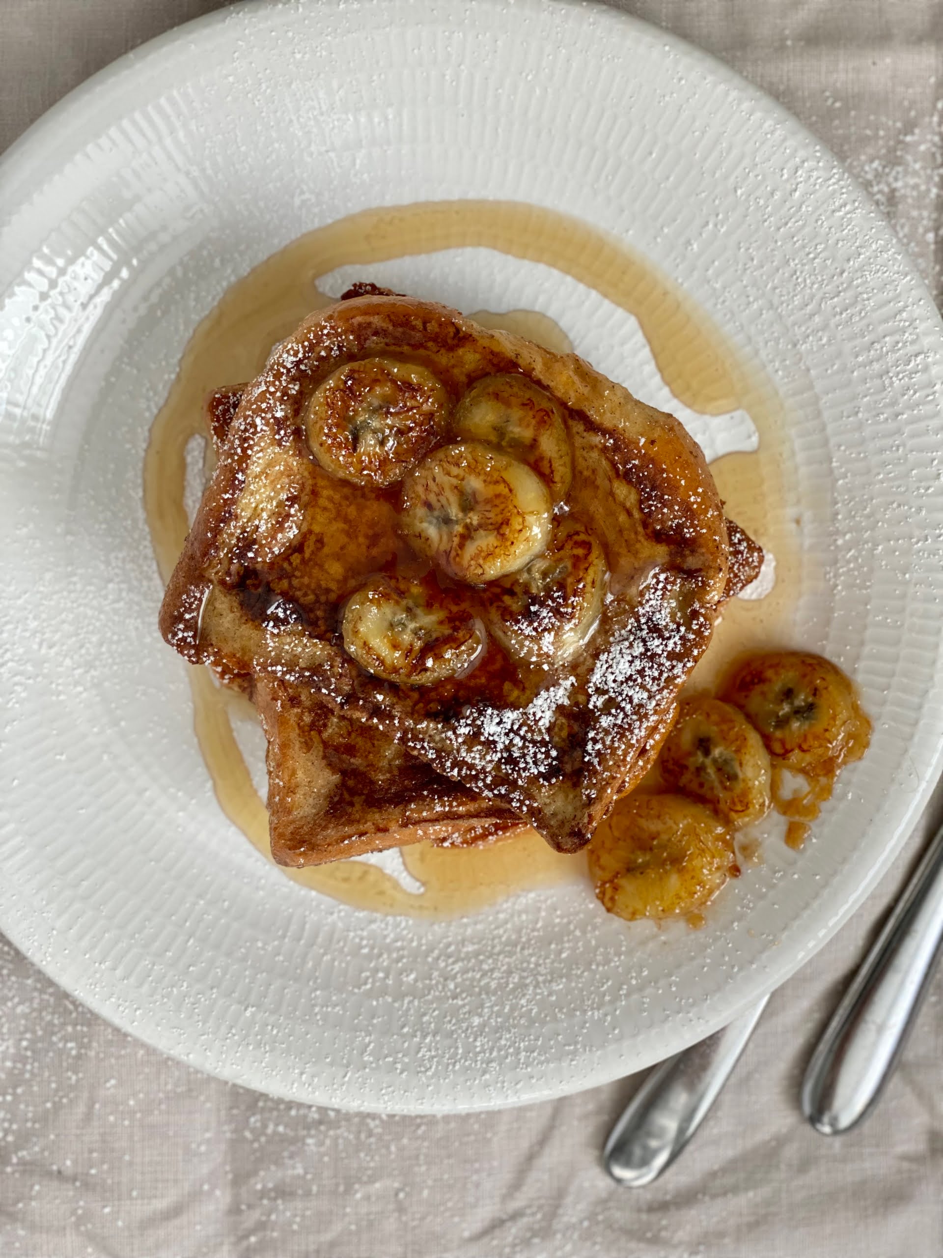 French Toast with Maple Caramelized Bananas on the top and 3 arranged on the side of the plate with a dusting of icing sugar and drizzled with maple syrup -Pure Maple