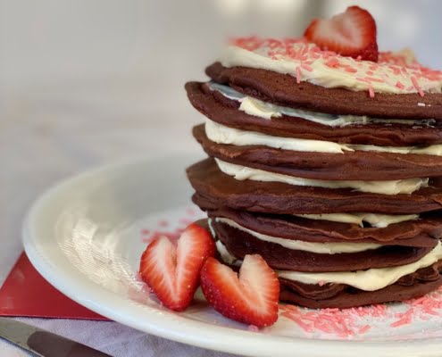 A stack of Red Velvet Pancakes with Maple Cream Cheese Frosting decorated with cut strawberries and pink sprinkles