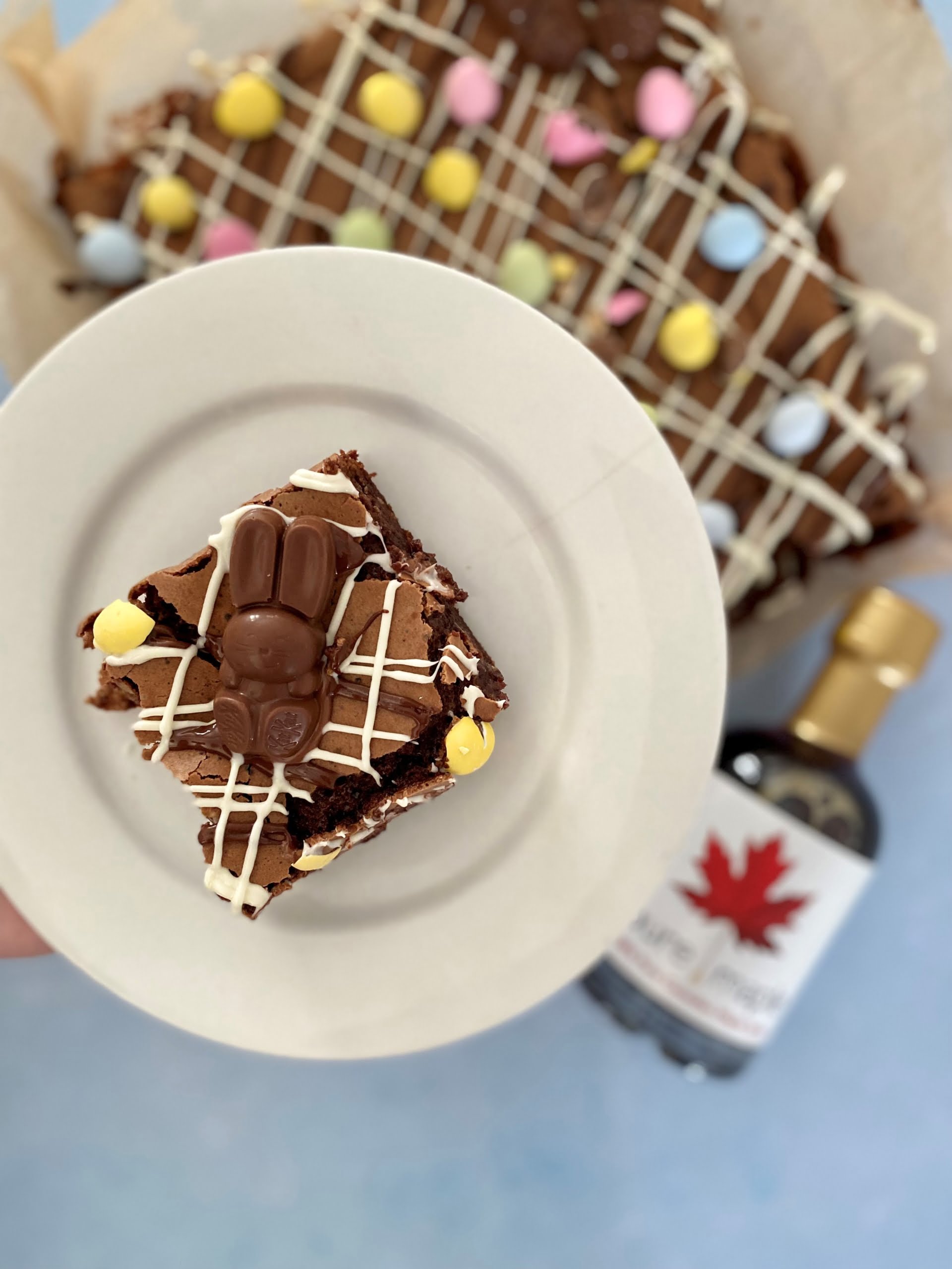A slice of Easter Egg Brownie on a plate with the brownie and maple syrup bottle in the background